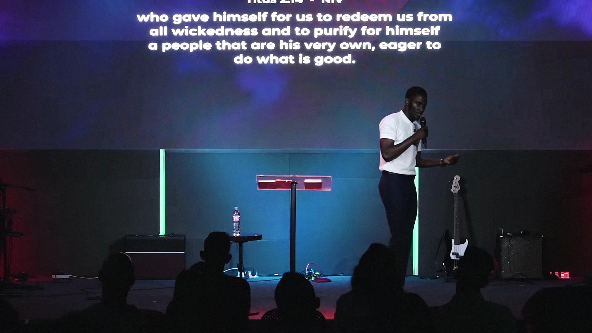 Alexander Hwande on a church stage, gesturing as he speaks, with a verse from Titus on the screen above him