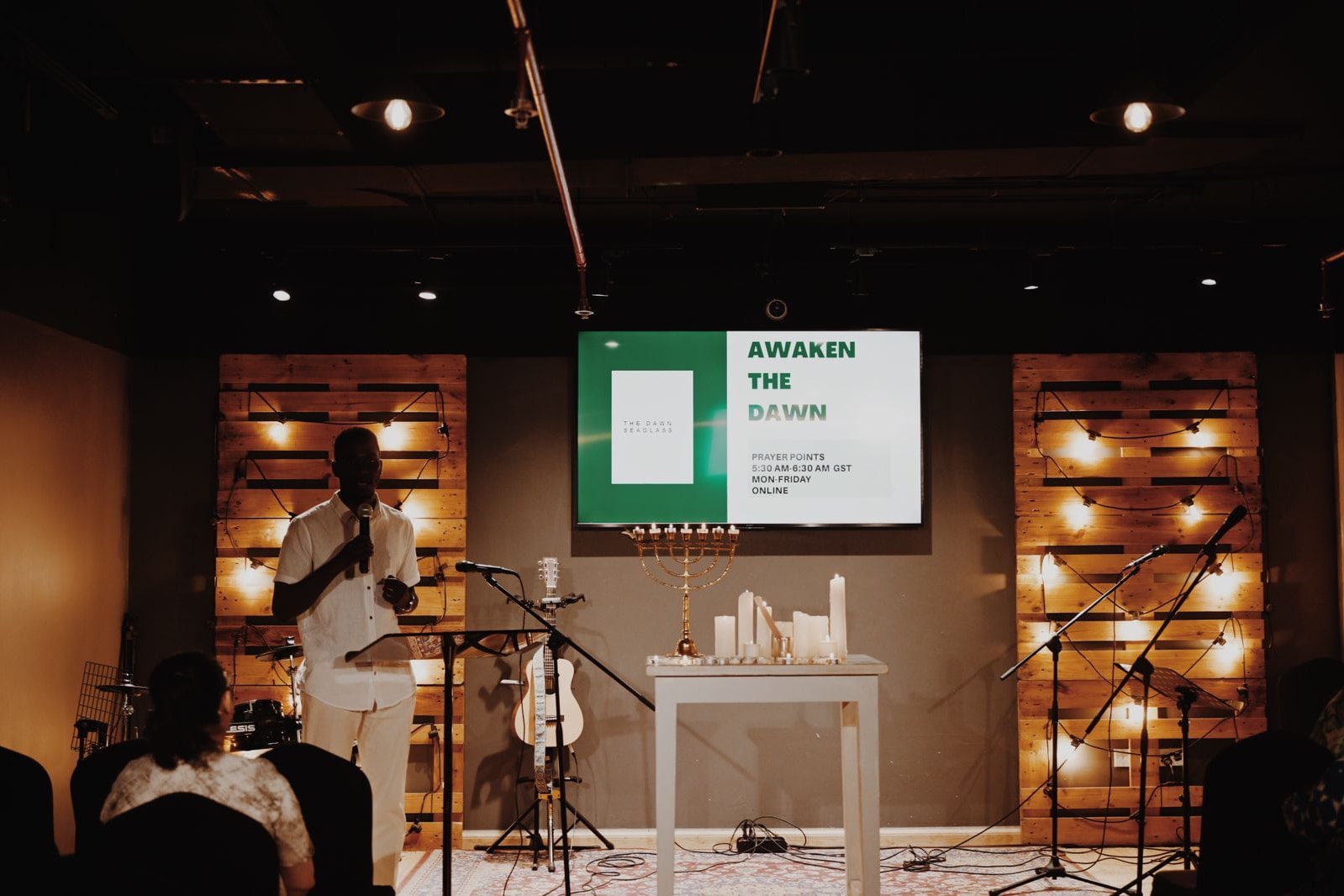 Alexander Hwande speaking at a prayer gathering, on a stage lit by strings of warm lights