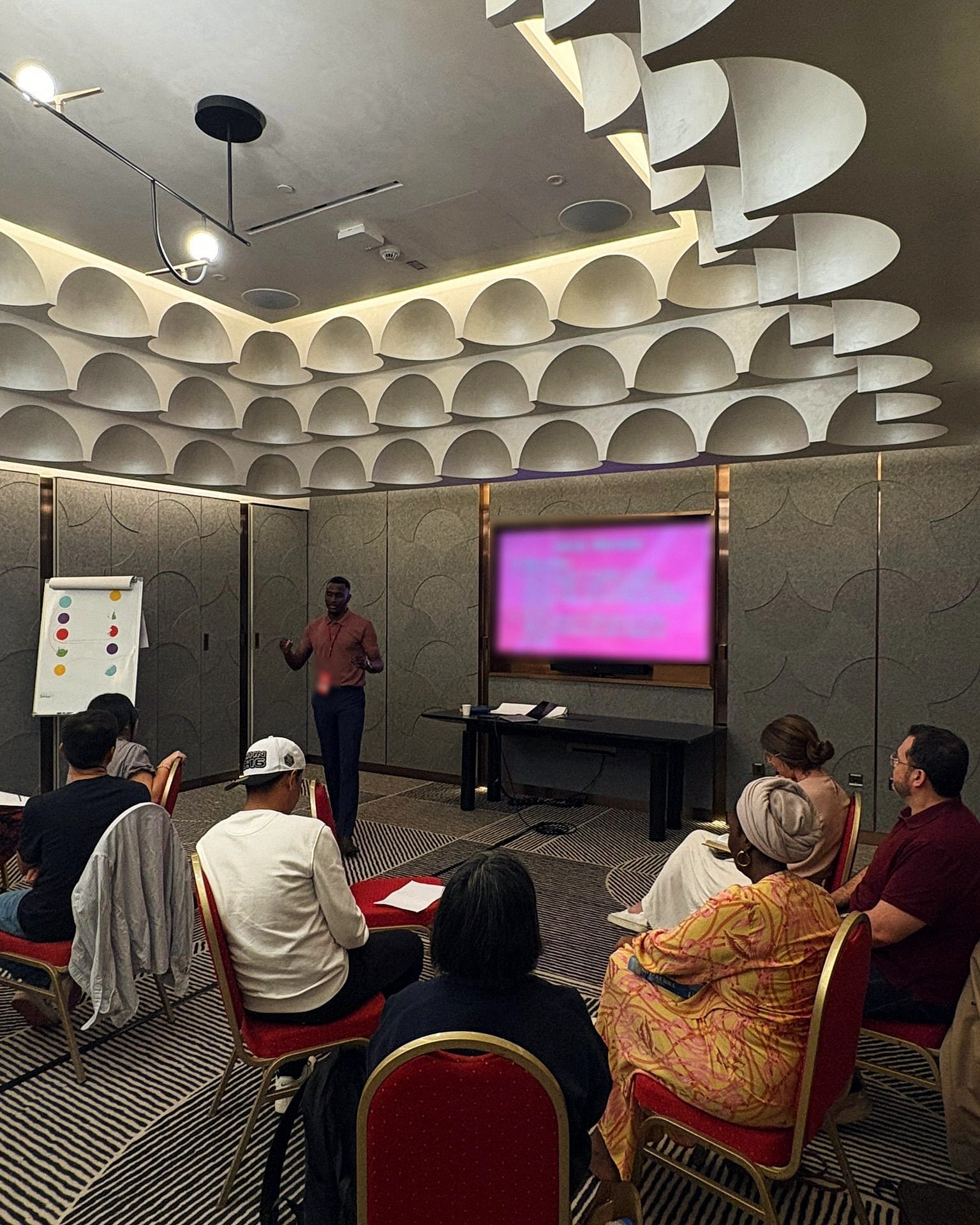 Alexander Hwande speaking to a seated group in a meeting room, beside a screen