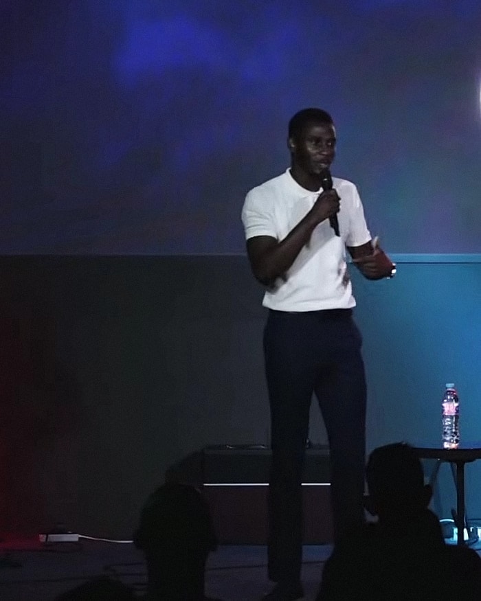 Alexander Hwande on a stage with a microphone, speaking to a seated audience