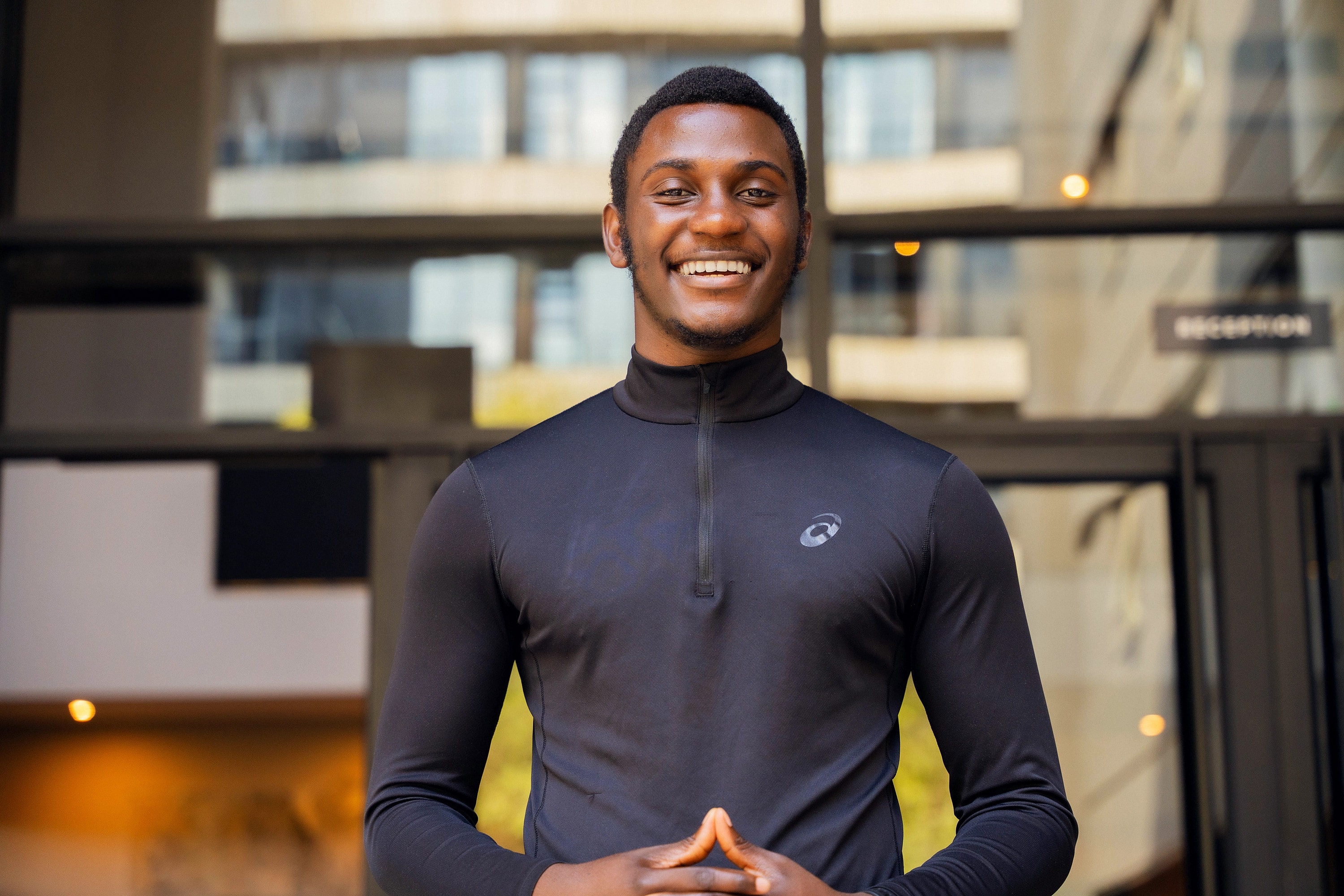 Alexander Hwande, smiling, in a black half-zip top with his hands together