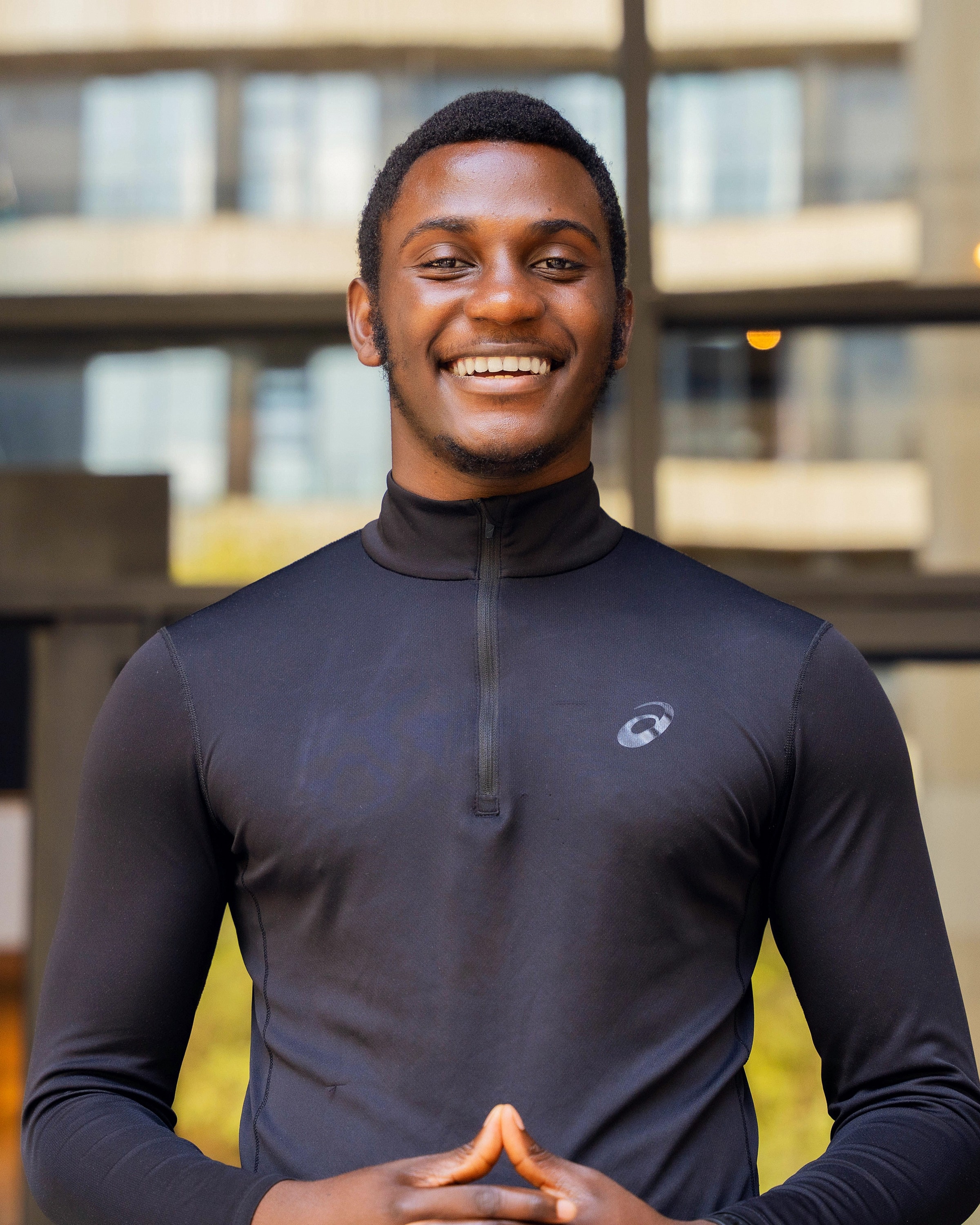 Alexander Hwande, smiling, in a black half-zip top with his hands together